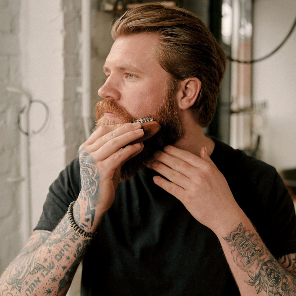 Man with tattoos grooming his beard with a Brooklyn Soap Company beard brush in a casual setting.