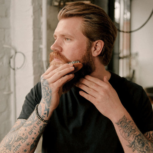Man with tattoos grooming his beard with a Brooklyn Soap Company beard brush in a casual setting.