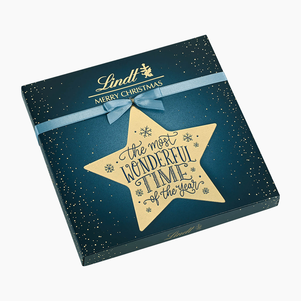 Lindt Merry Christmas Calligraphy chocolate pralines box in dark teal blue with gold speckles, a light blue ribbon, and a large gold star reading 'the most wonderful time of the year'.