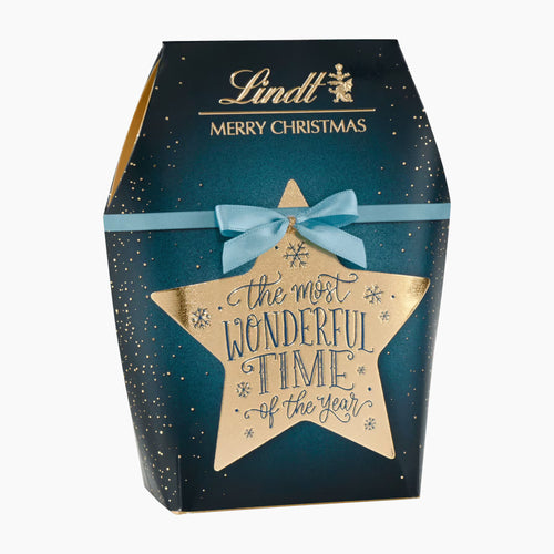Lindt Merry Christmas Calligraphy XMAS pralines bag gift box with a dark teal-blue background, gold glitter speckles, a gold star-shaped front label reading 'the most wonderful time of the year,' a light blue ribbon bow, and gold Lindt branding at the top.