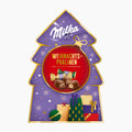A Christmas tree-shaped Milka Christmas Pralines chocolate gift box with a purple background, gold trim, a red circular center, and holiday gift illustrations. The Milka logo appears at the top, and the center text reads 'Weihnachtspralinen'.