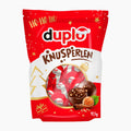A red and white bag of Duplo Knusperlen holiday chocolate pralines, featuring illustrations of chocolate pralines, hazelnuts, and festive gold stars on a white background.