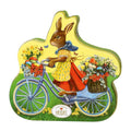 Heidel Easter Nostalgia Gift Tin Pralines with a Vintage-style Easter bunny on a bicycle with flowers, wearing a yellow dress and red skirt, on a white background.