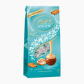 A package of Lindt Lindor Cinnamon Roll limited edition chocolate pralines, featuring a turquoise blue bag with gold and white decorative elements, a large chocolate cinnamon roll, and smaller wrapped chocolates.