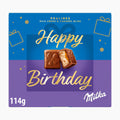 A square Milka chocolate pralines box with a blue and purple design and gold 'Happy Birthday' lettering, featuring two milk chocolate pralines on the front and '114g' in the lower left.